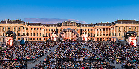 Besucherreihen die bei einer Veranstaltung im Schlosspark vor dem beleuchteten Schloss Schönbrunn sitzen 
