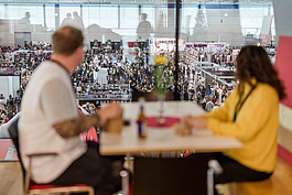 Zwei Besucher die an einem Tisch sitzen und durch eine Glasscheibe auf ein Veranstaltung in der Messe Wien blicken